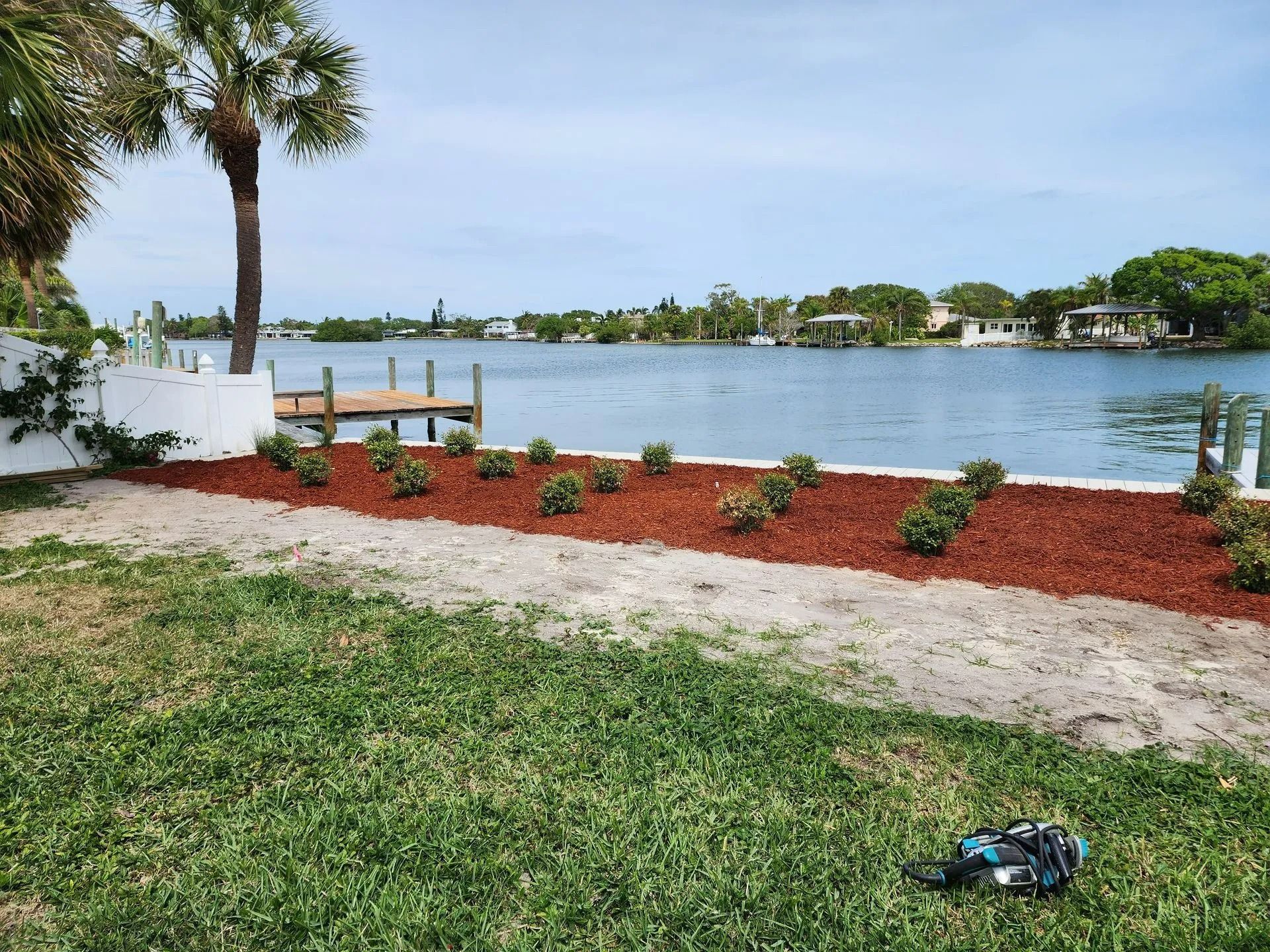 Lawn and landscaped yard border a waterway with a dock, white wall, and homes visible in the distance.