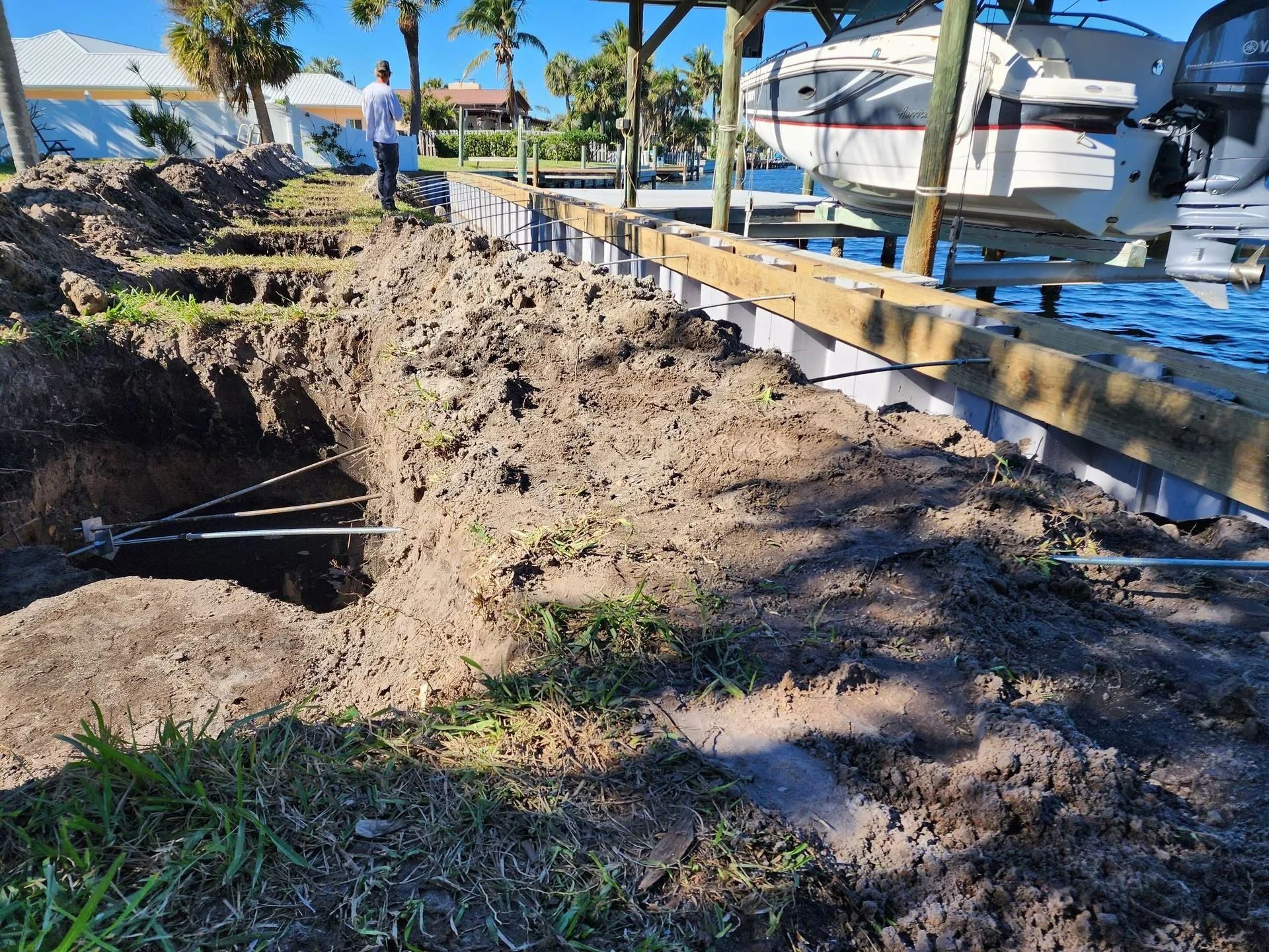 Construction site with a dug-out trench, soil, and wooden support beams near a boat dock and waterway. A person is walking in the background.