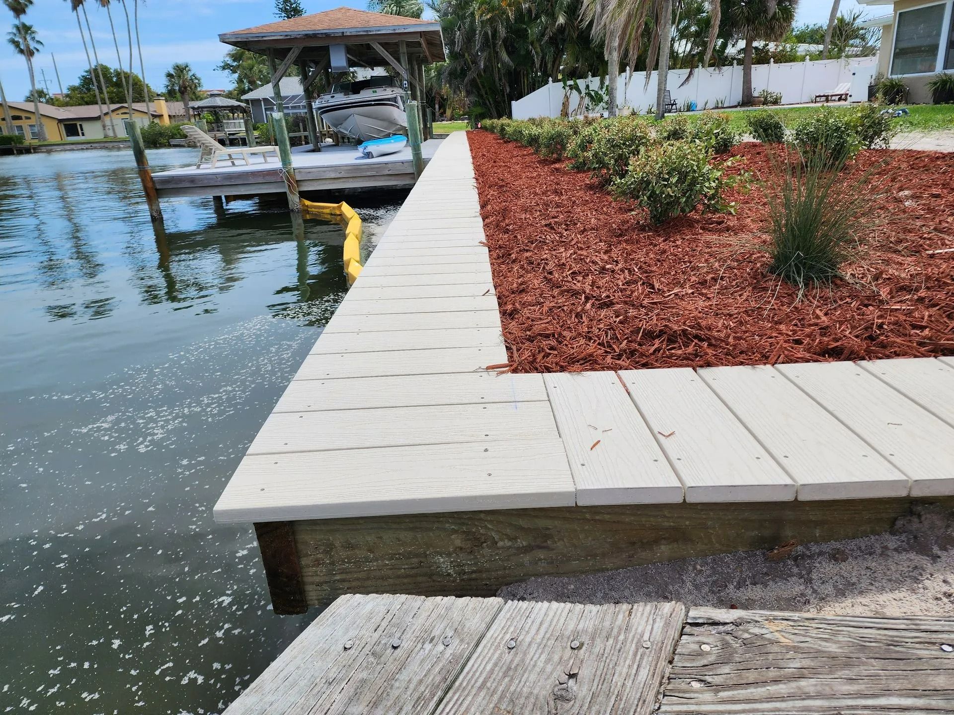 A waterfront with a light-colored composite retaining wall, a red-brown mulched garden bed, and a wooden dock.