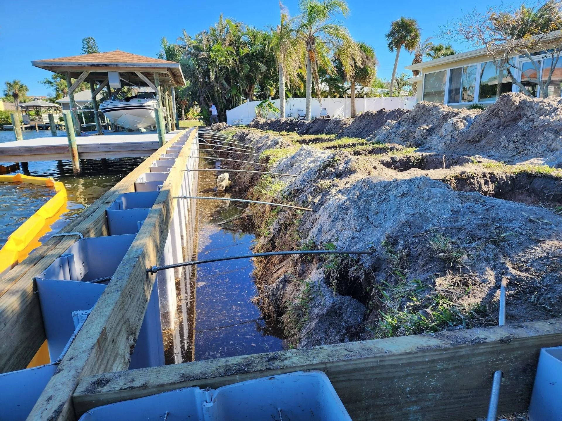 Construction site with a waterfront retaining wall being built; dirt piles and water are visible. Wooden beams and metal bars are in place.