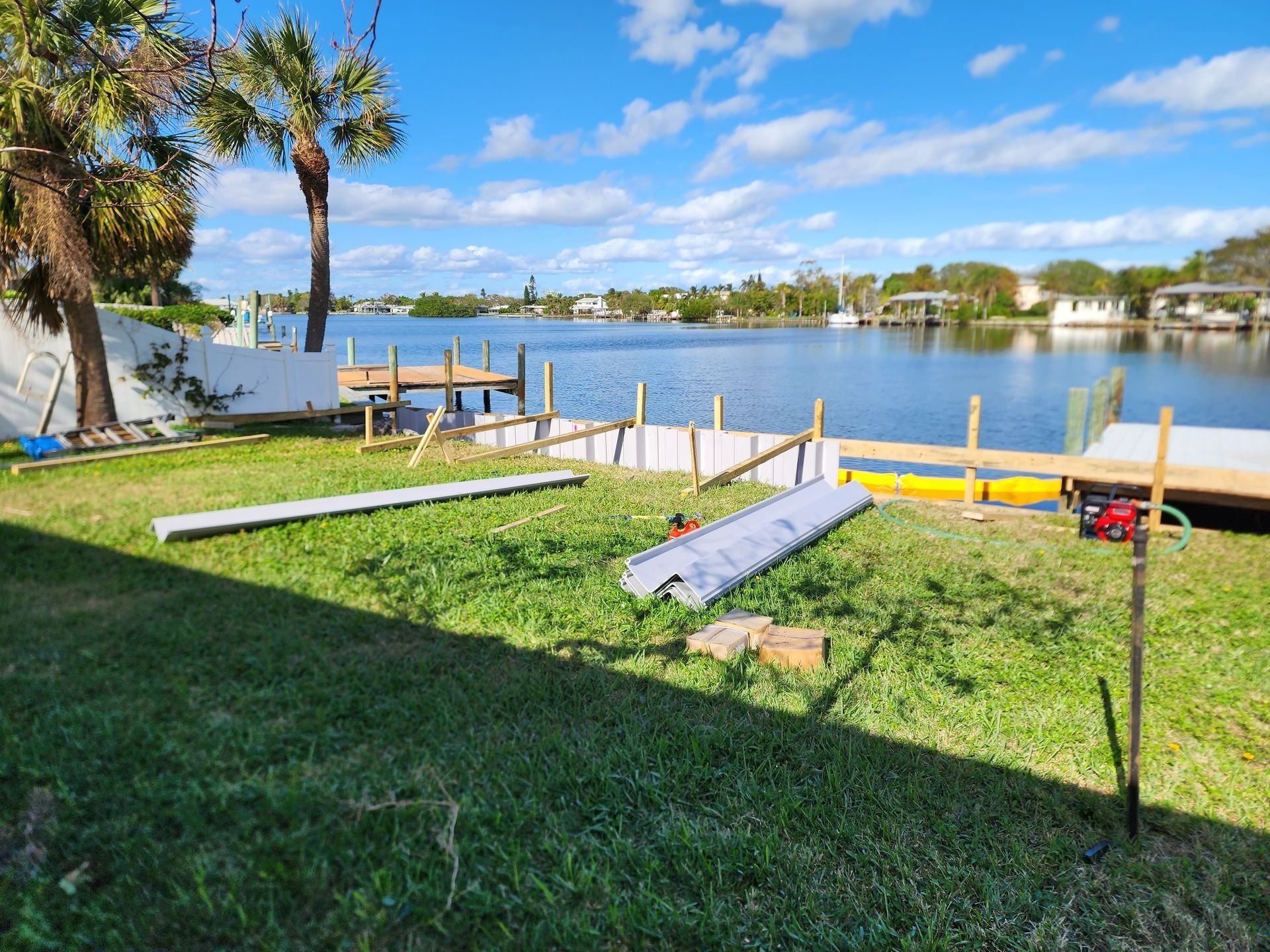 Construction in a yard by a waterway. Boards and equipment are on the grass, leading to a dock. Houses and blue sky are in the background.