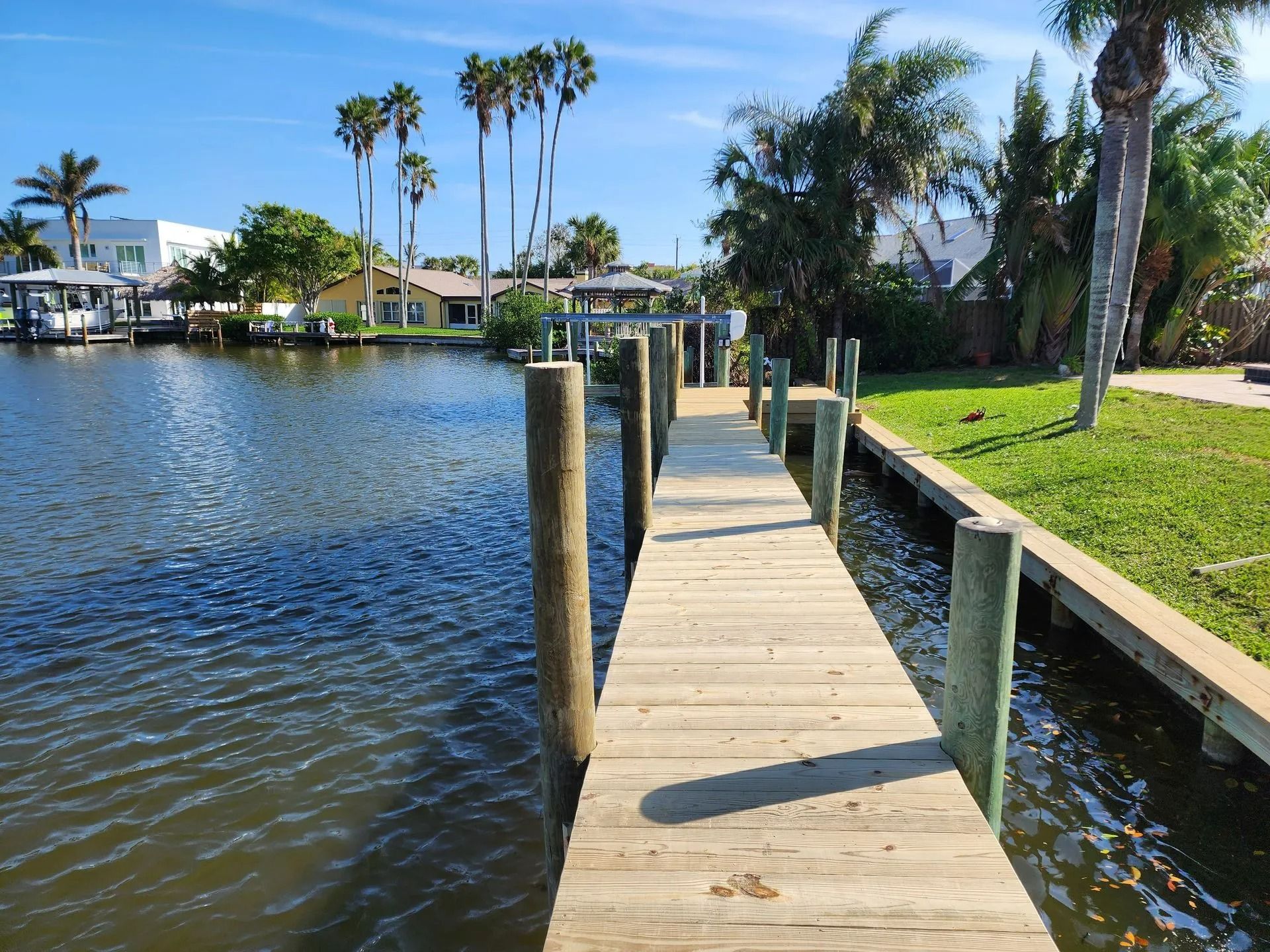 Wooden dock extending into a canal, flanked by wooden pilings. Palm trees and houses are visible in the sunny background.