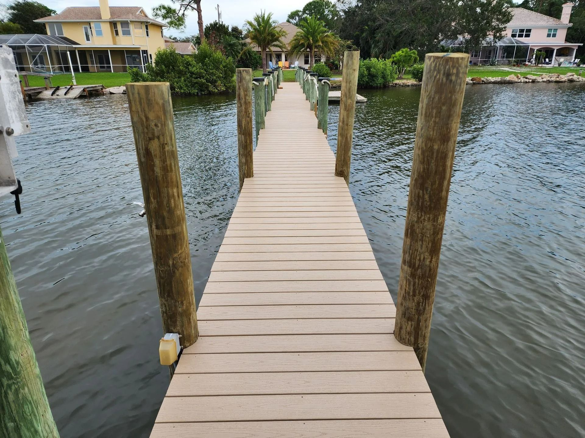 Wooden dock extending into a waterway, bordered by pilings. Houses and greenery are visible in the background.