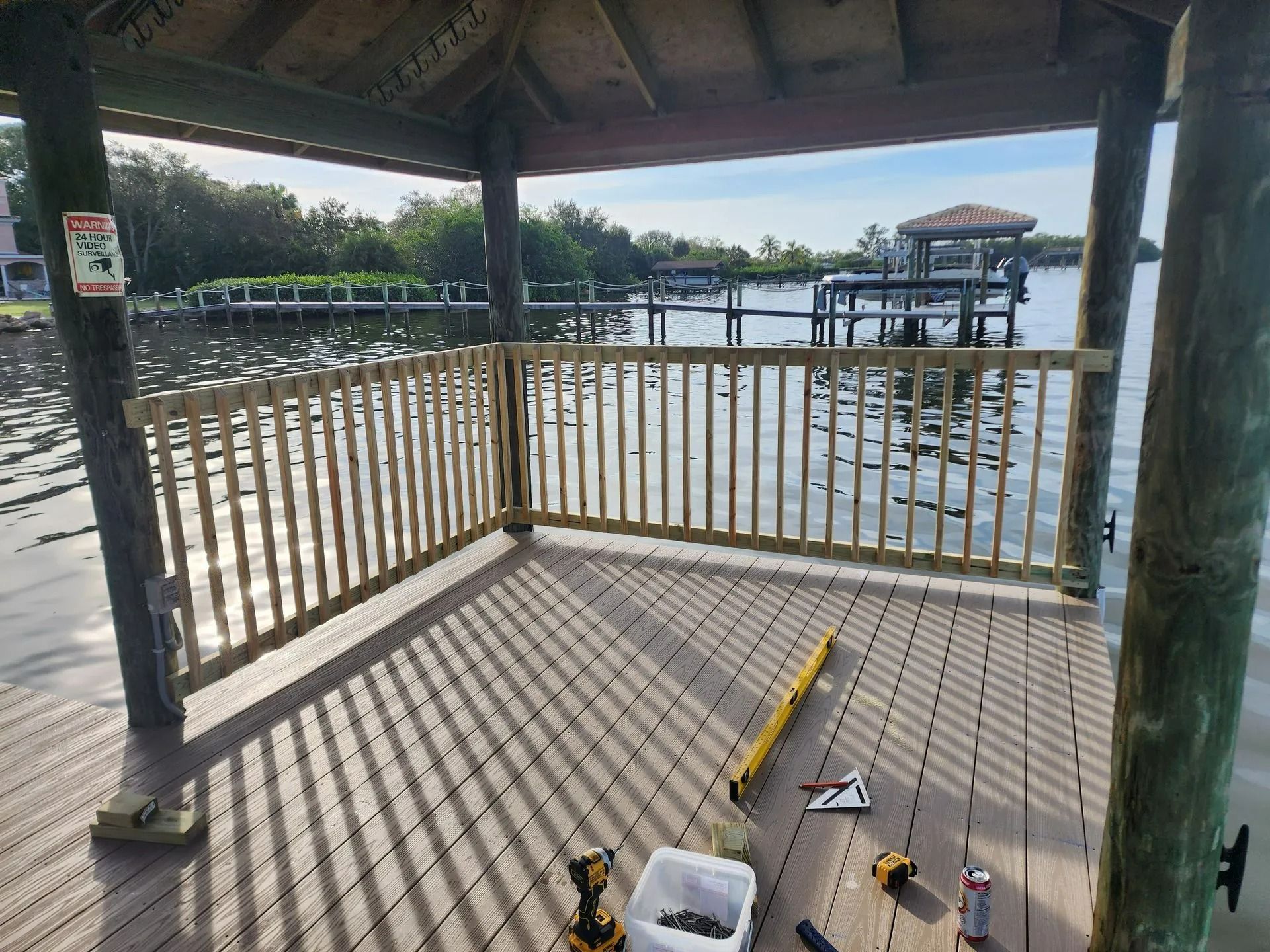 A wooden dock with a gazebo-like structure and railing overlooking a waterway. Tools and a bucket are visible on the deck.