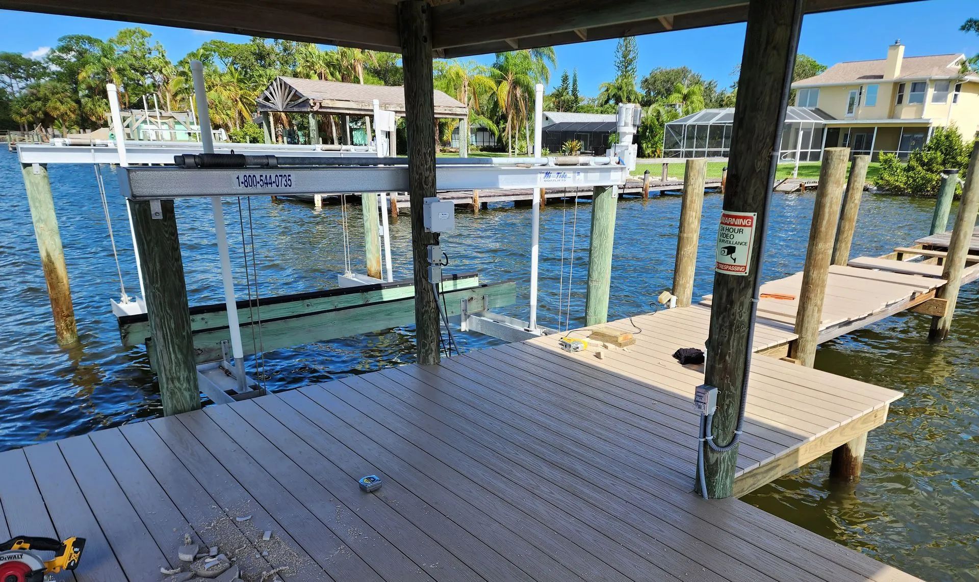 Dock with boat lift over water, wooden deck, surrounding houses and trees under a blue sky.