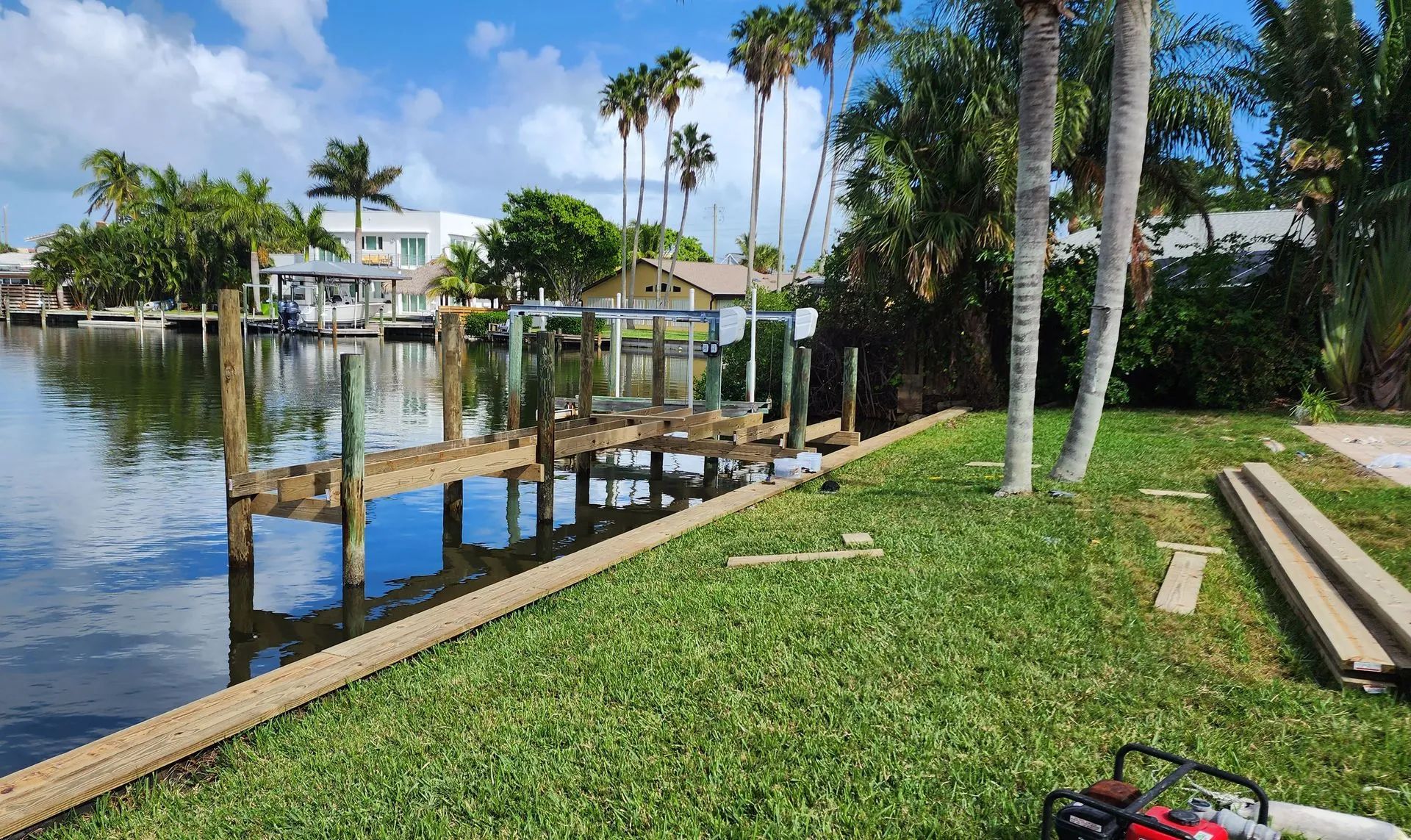 Dock on a canal with a boat lift, surrounded by green grass, palm trees, and houses under a blue sky.