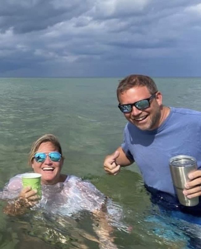 Two people smiling in shallow ocean water under cloudy skies, each holding a drink.