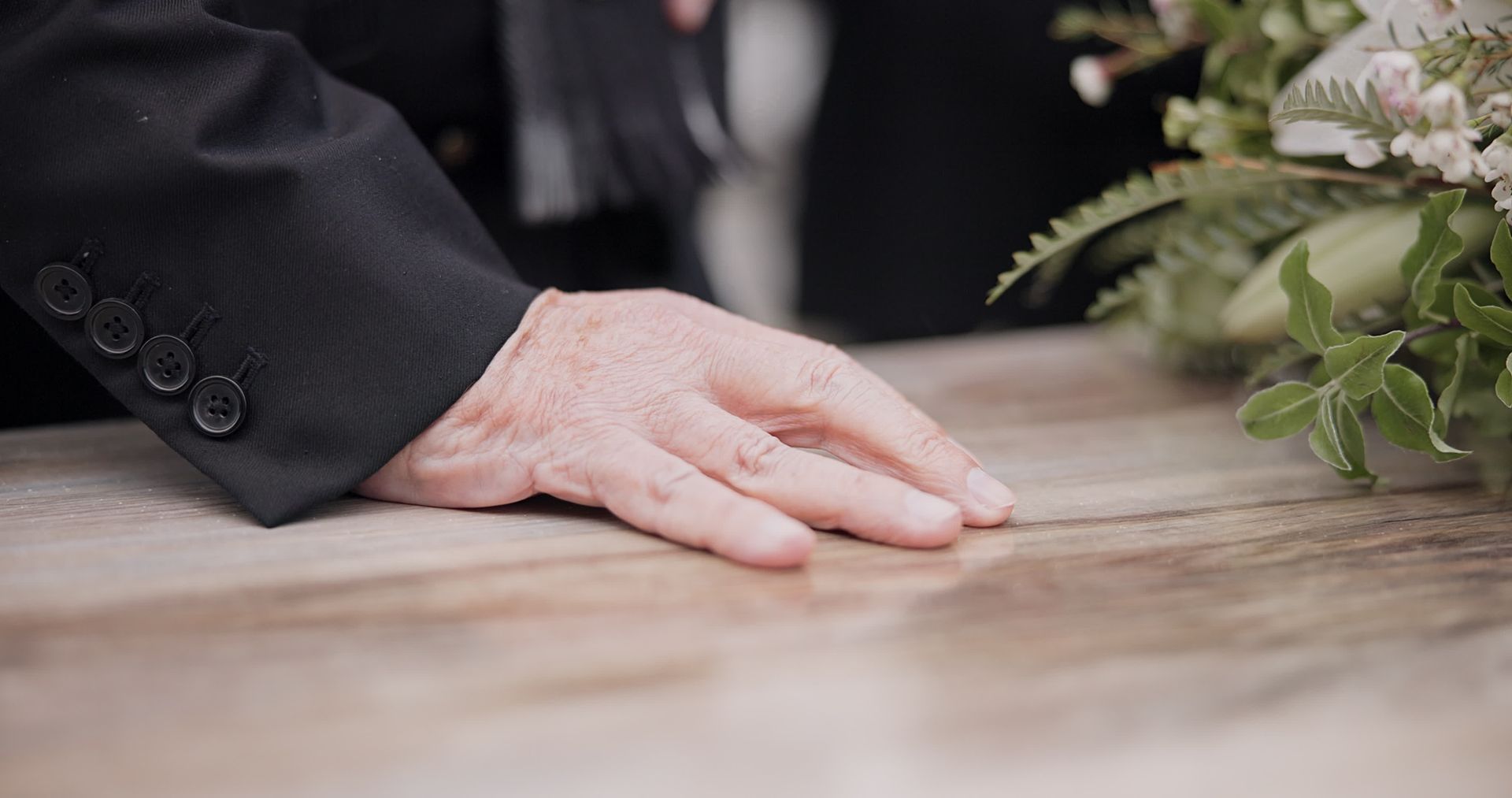 A hand rests gently on a wooden casket adorned with flowers at a funeral service.