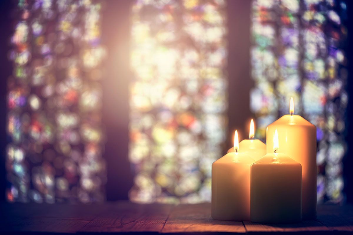 Lit candles on a wooden surface in front of a colorful stained-glass window.