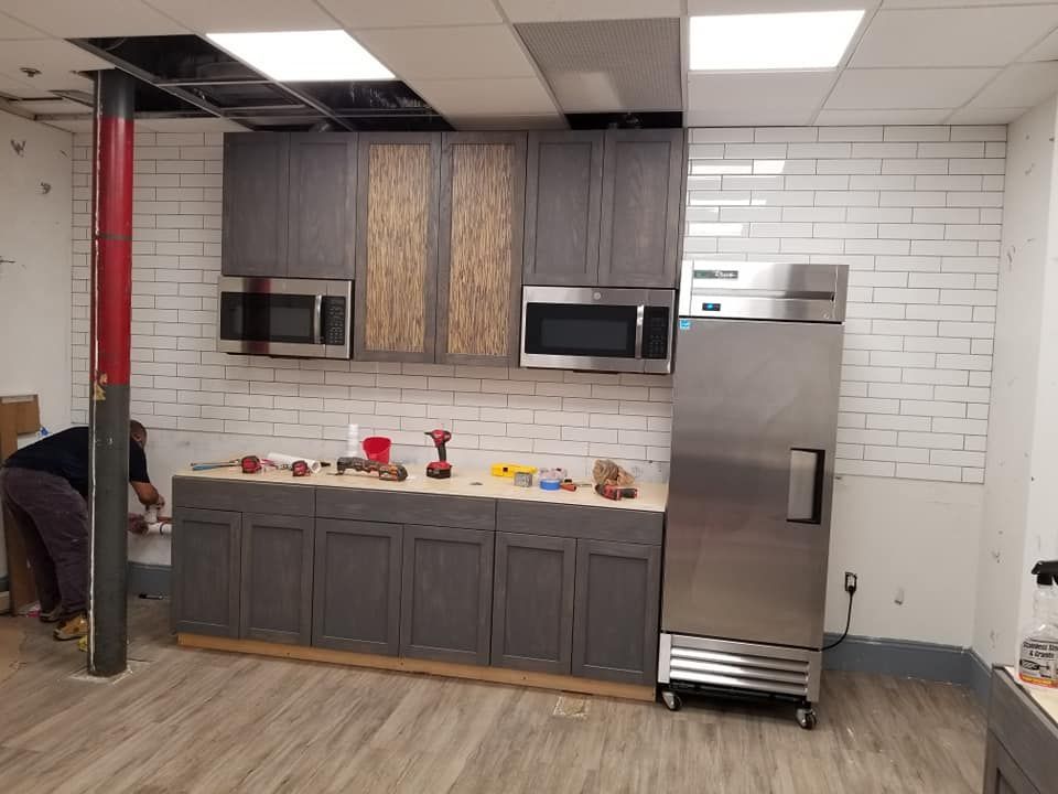 A person installs cabinets in a kitchen with a stainless steel refrigerator, white brick wall, and gray cabinets.