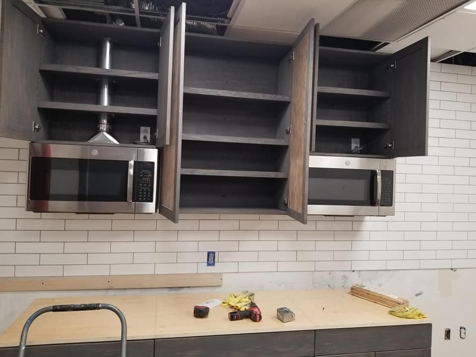 Kitchen with gray cabinets, two microwaves, and shelves.  A counter and brick-patterned wall are below.