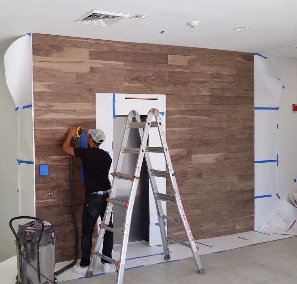 Man using power tool on wood-look tile wall, standing on ladder. Construction in progress.