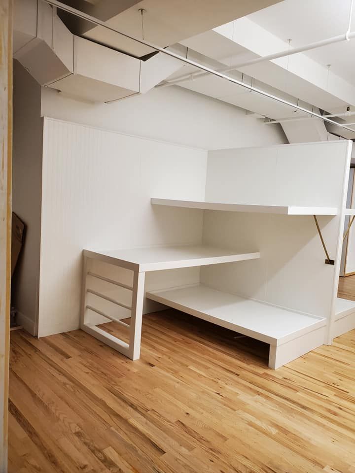 White built-in desk and shelves in a corner against a white wall, set on a wooden floor.