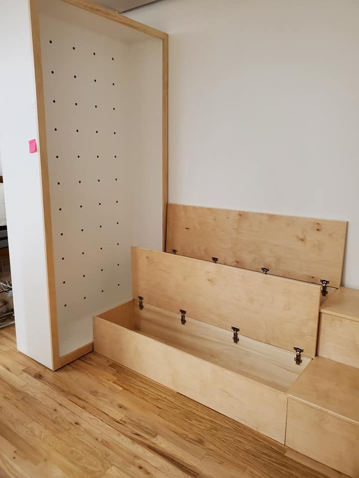 Wooden built-in storage unit against a white wall. Open compartments with hinged lids and a tall, slotted cabinet.