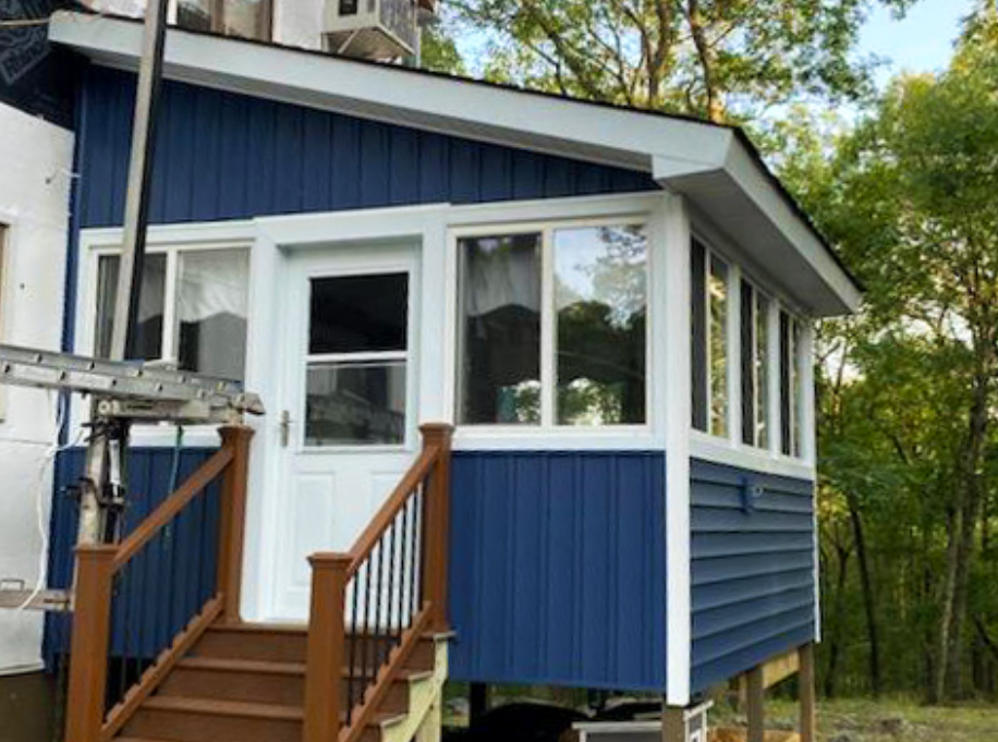 Blue-sided porch with white trim and door, brown stairs, and forest backdrop.