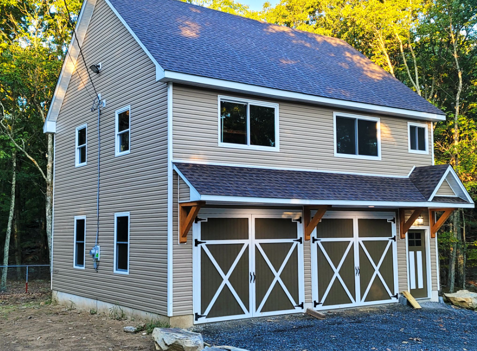 Two-story house with tan siding and brown garage doors; a dark blue roof, set in a wooded area.