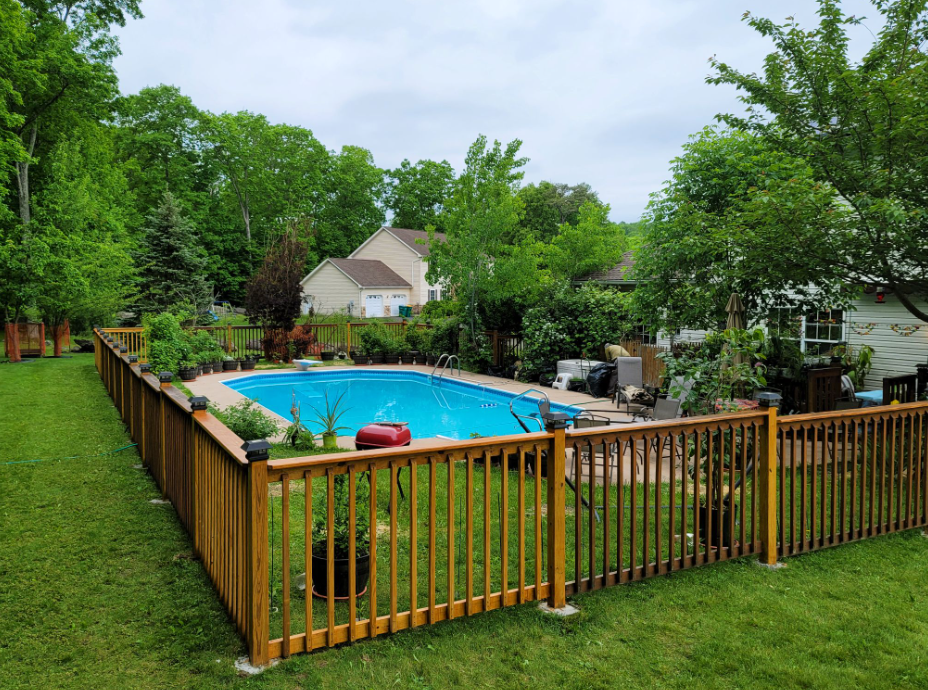 Wooden fence surrounds a backyard swimming pool. Green trees and lawn surround the pool with a cloudy sky above.