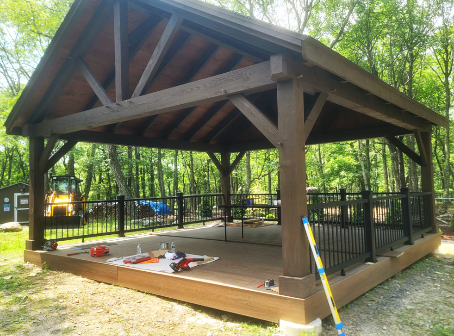 Wooden pavilion with brown beams, dark railings, and a composite deck in a wooded area.