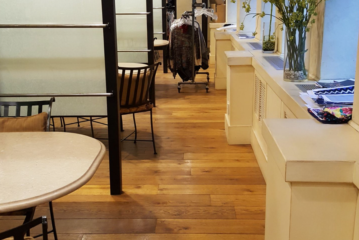 Wooden floor in a store, with tables, chairs, clothing racks, and windows with sunlight.