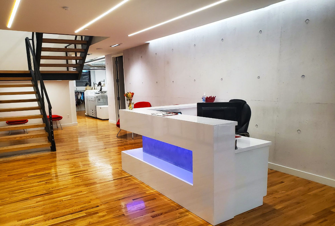 Modern office reception area with white desk, wooden floor, and stairs.