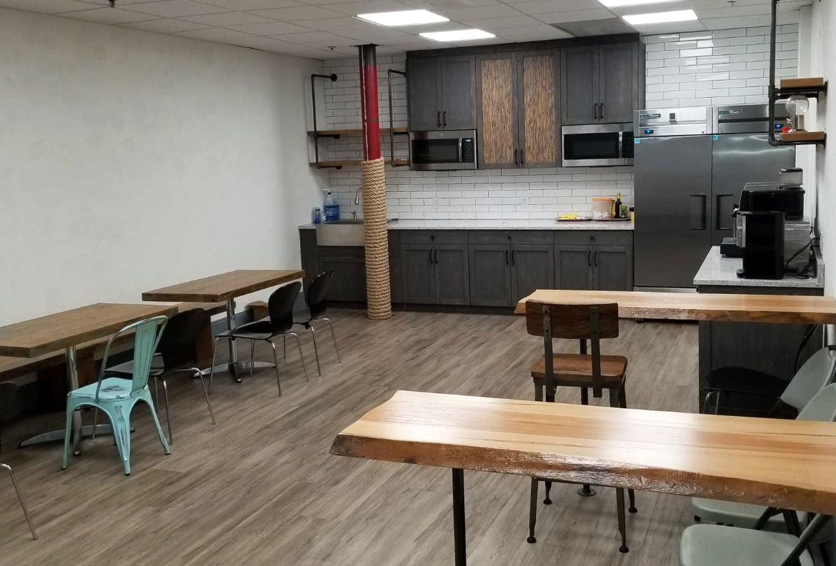 Cafeteria with tables, chairs, and a kitchen. Dark cabinets, white tile backsplash, and wood and gray tones.