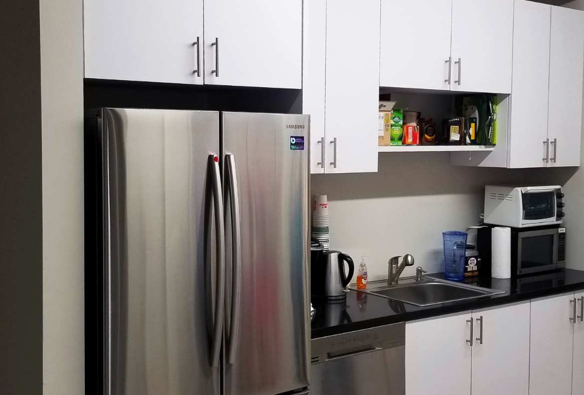 Stainless steel refrigerator beside white kitchen cabinets, countertop, sink, and microwave.