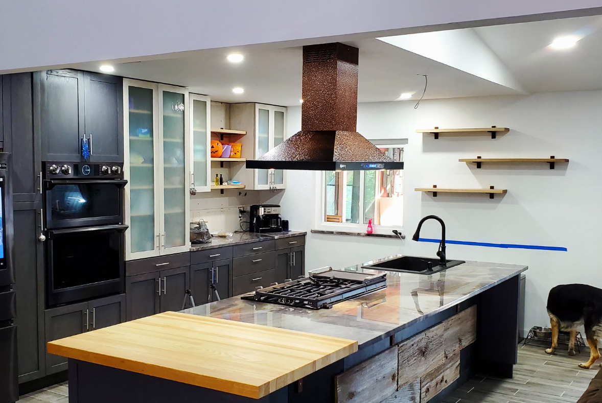 Modern kitchen with island, oven, range, copper hood. Dark cabinets, butcher block, and floating shelves.