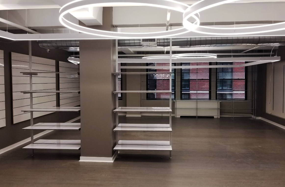 Empty retail space with shelving, a brown support column, and windows. Bright, circular lights illuminate the ceiling.