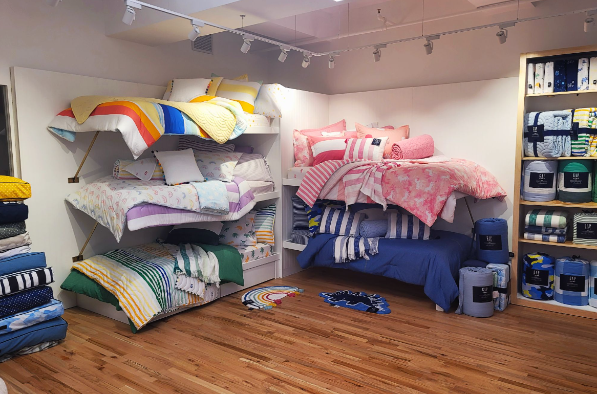 Beds with colorful bedding on display in a store, including shelves of folded blankets and pillows.