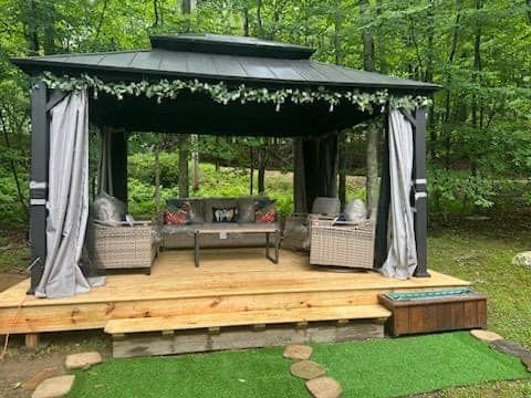 Gazebo with seating on a wooden deck, surrounded by greenery. Includes furniture and hanging curtains.
