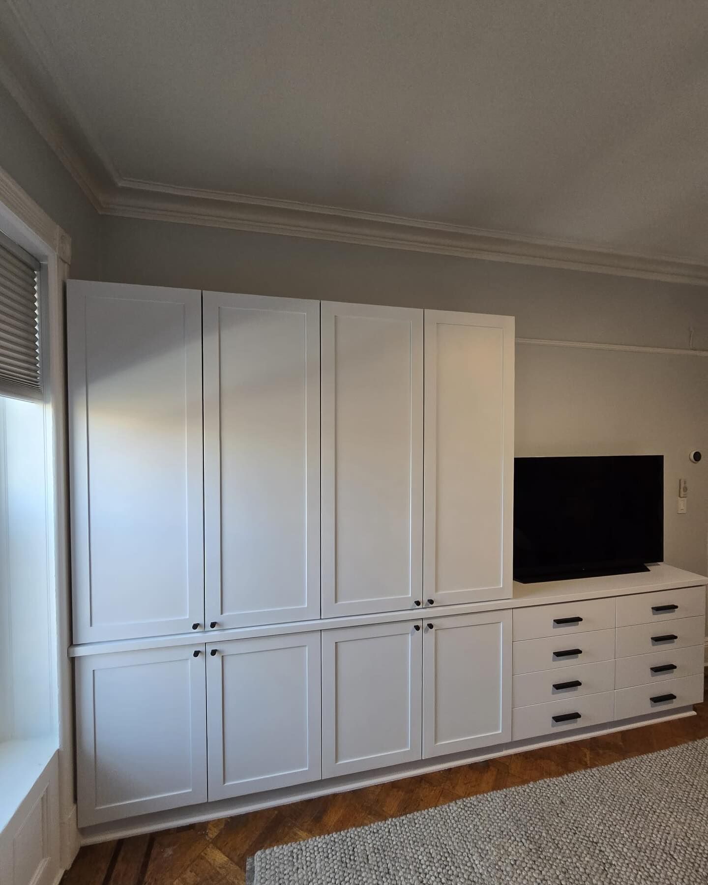 White built-in cabinets and drawers with a TV on top, set in a room with a rug and window.