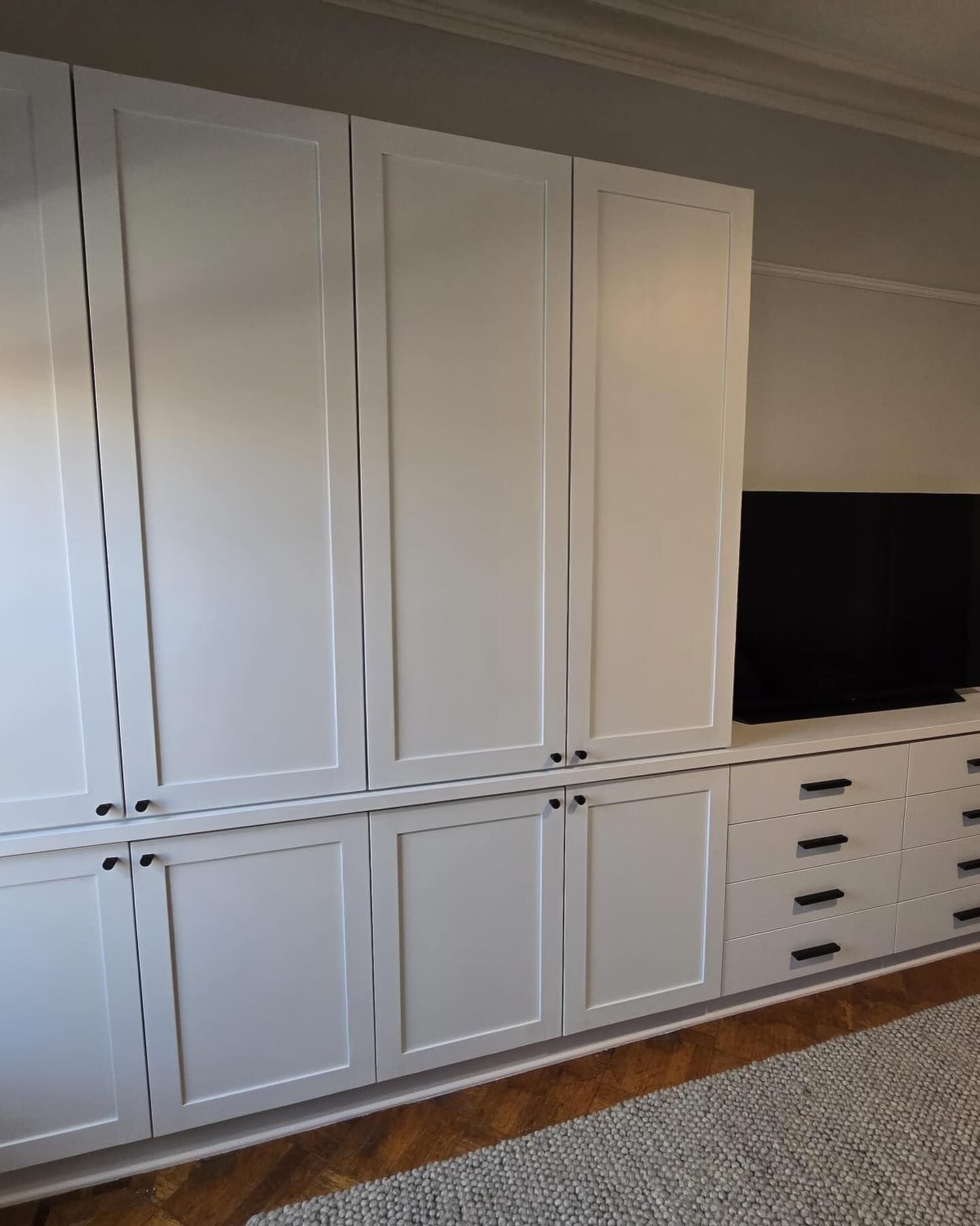 White built-in cabinet with black handles, a TV, and drawers.  Wooden floors with a gray carpet.