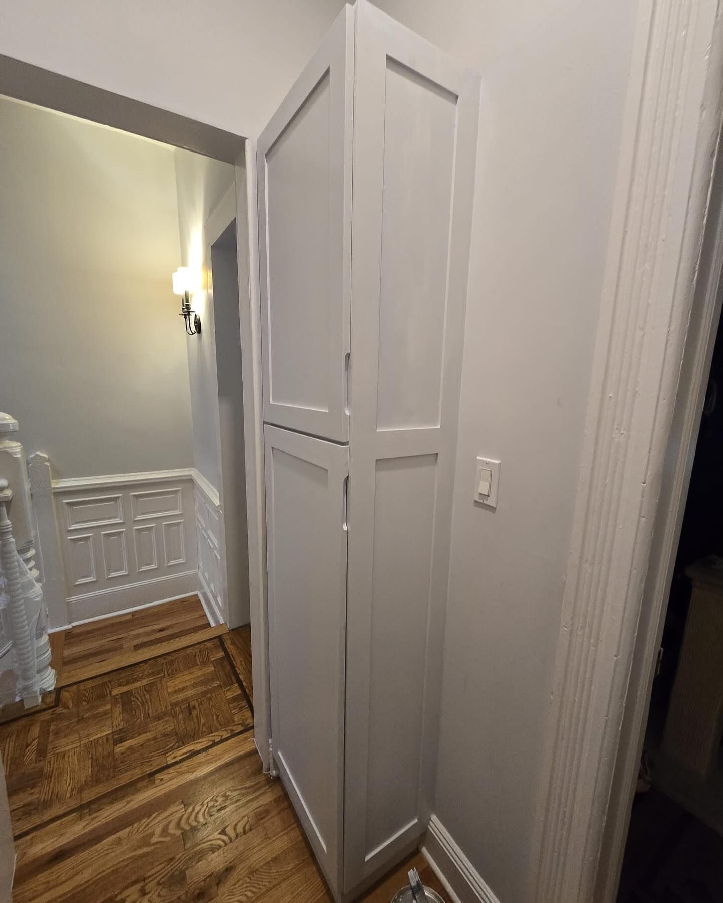 Tall, white cabinet in a hallway with wood flooring and a light switch.