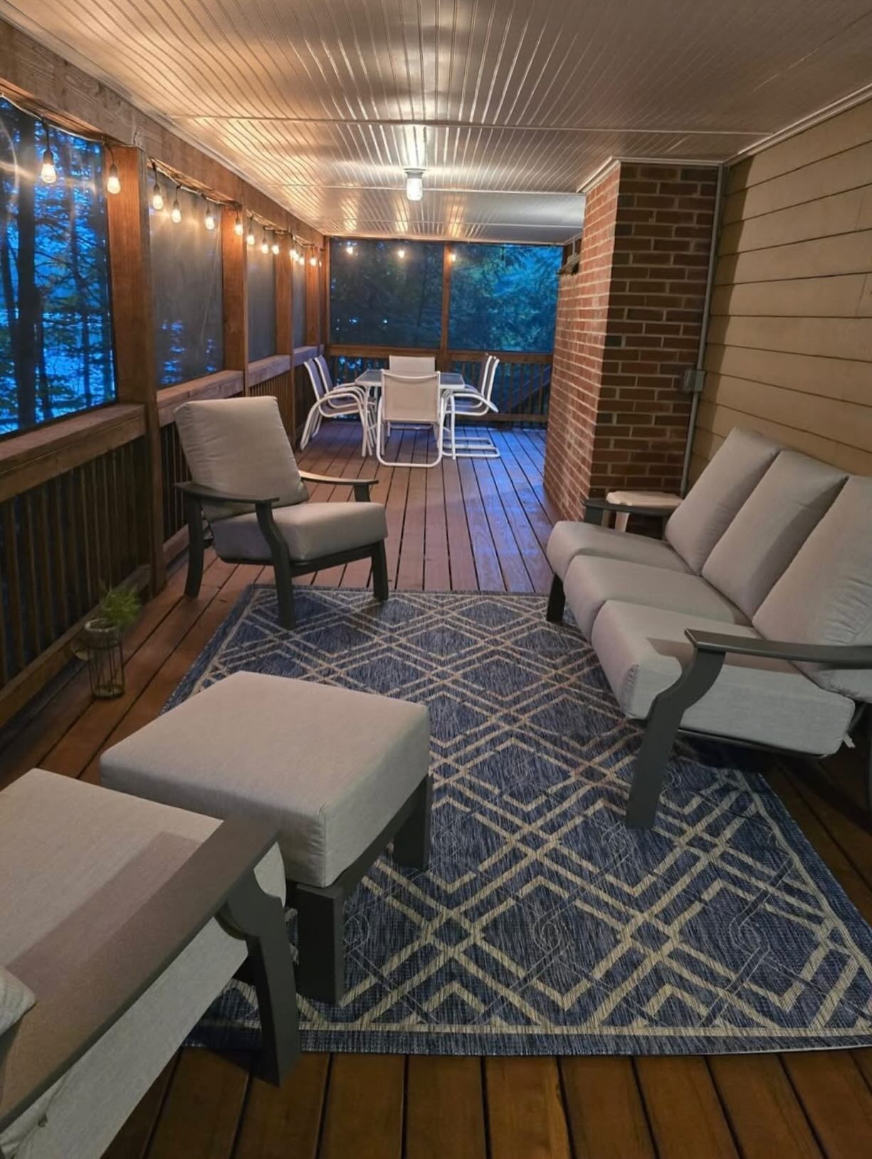 Screened porch with wooden floor and ceiling. Blue rug, light furniture, string lights, and a table.