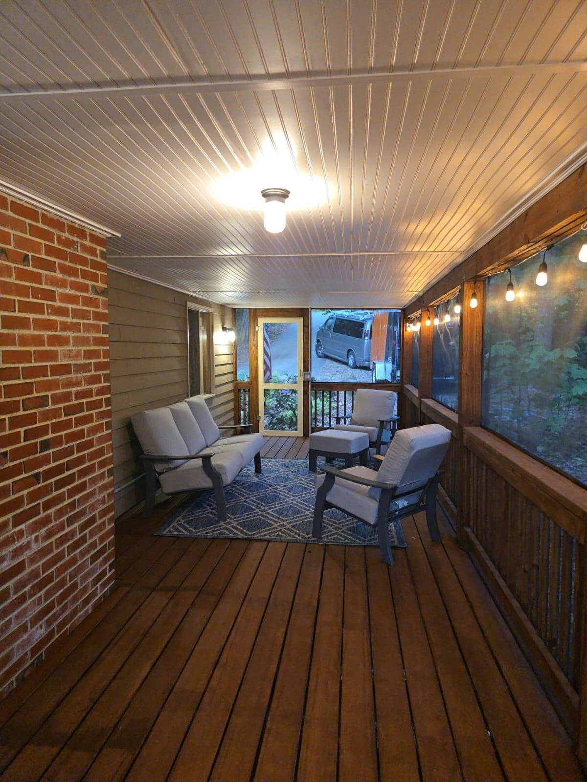 A furnished covered porch with seating and string lights, next to a brick wall.