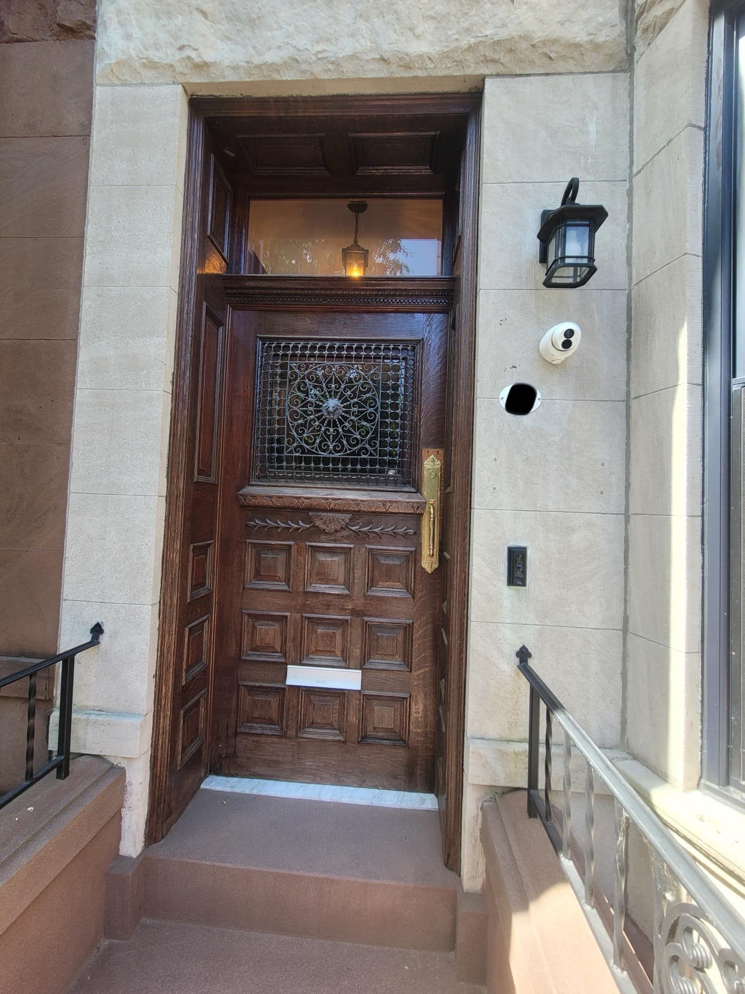 Ornate wooden door with decorative ironwork, brass handle, flanked by stone trim, small porch, and security camera.