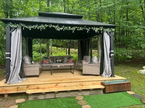 Gazebo with seating, wooden deck, curtains.  Set in wooded area.