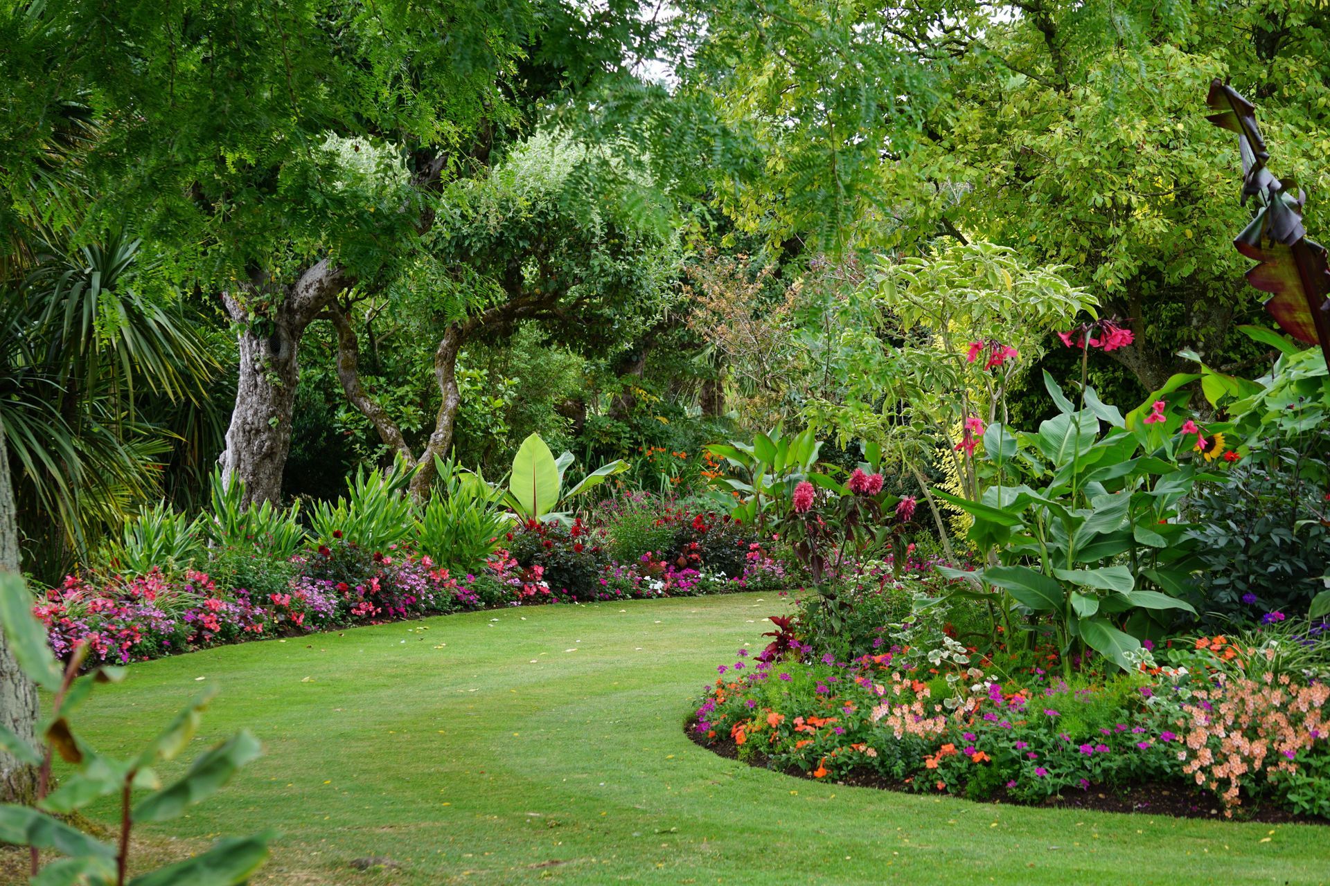 Lush green garden with a manicured lawn, dense shrubs, and purple flowers under trees