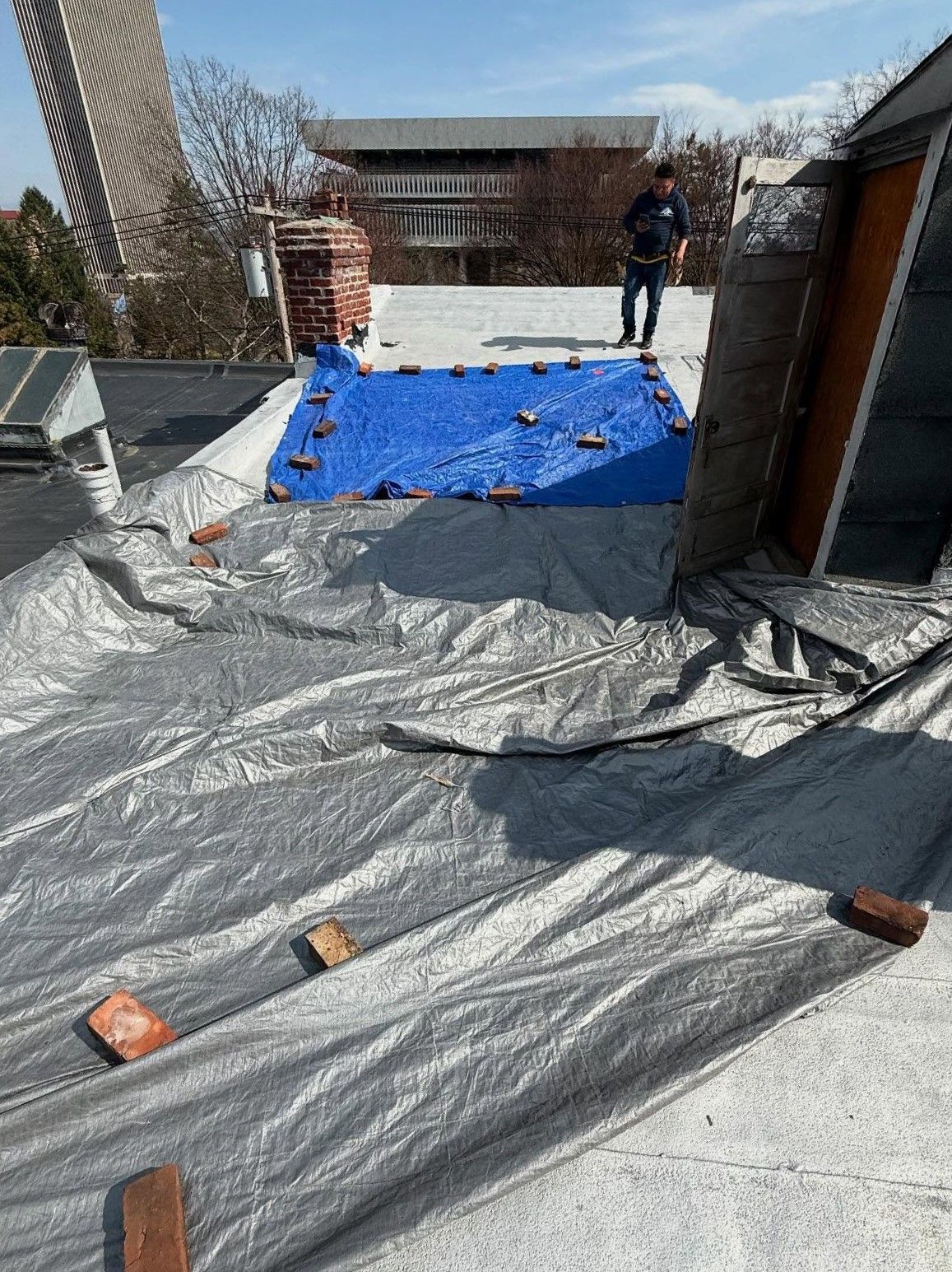 A man is standing on a roof with a tarp and bricks on it.