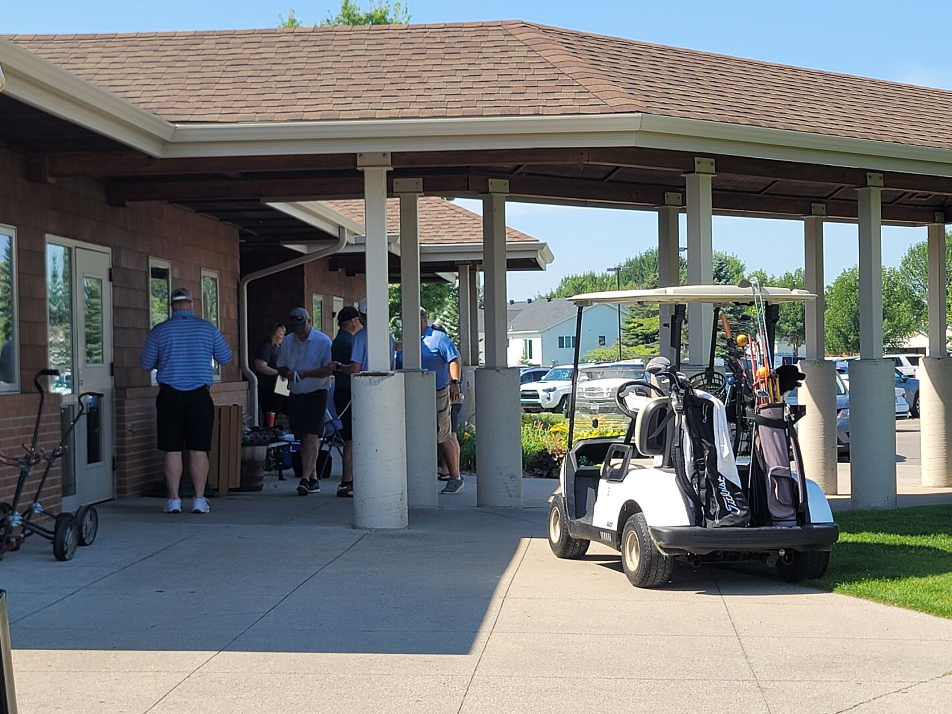 Golfers near a clubhouse with golf carts and bags, under a covered area, outdoors.