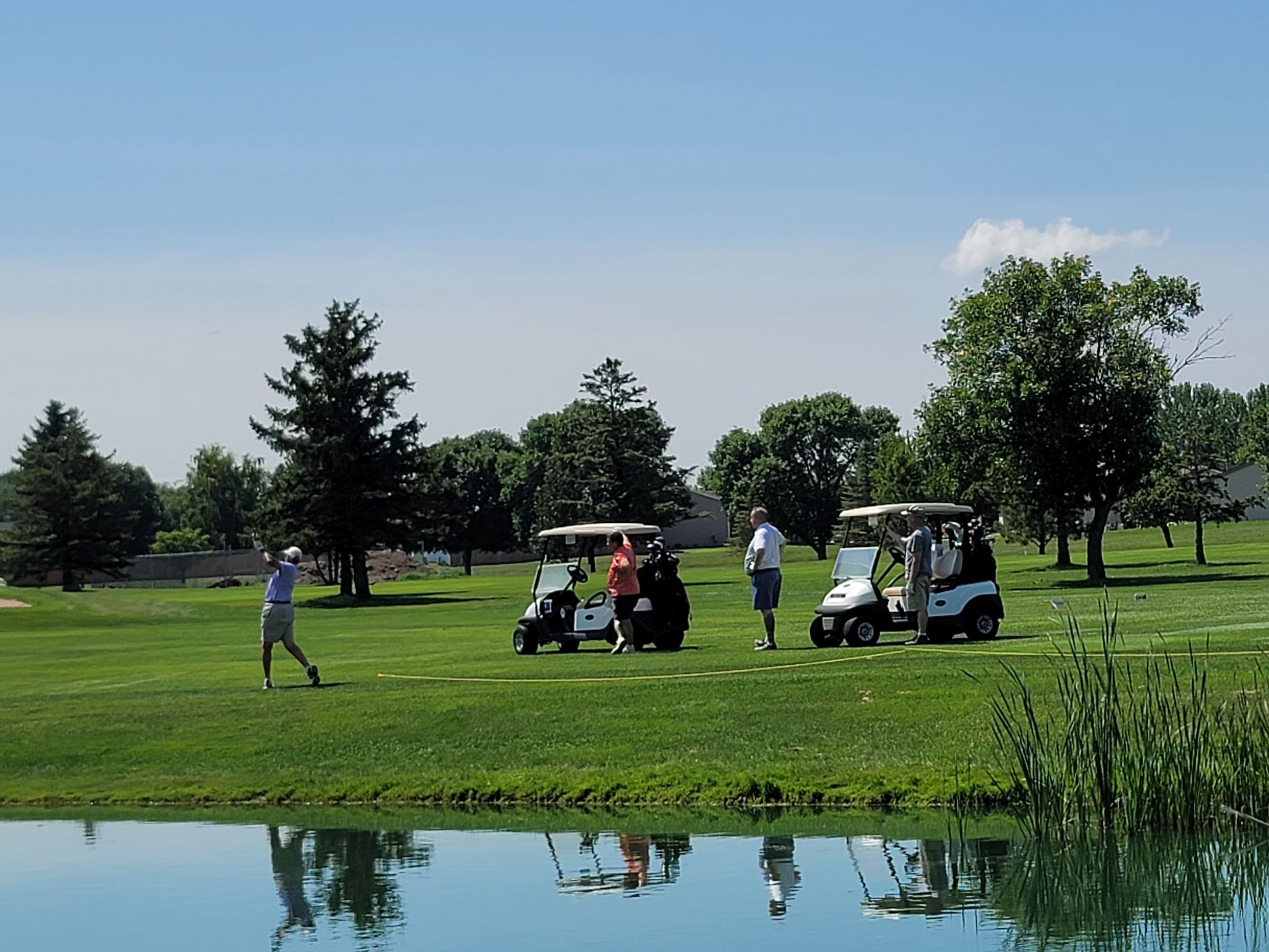Golfer swings on a green golf course near water with golf carts and people on a sunny day.