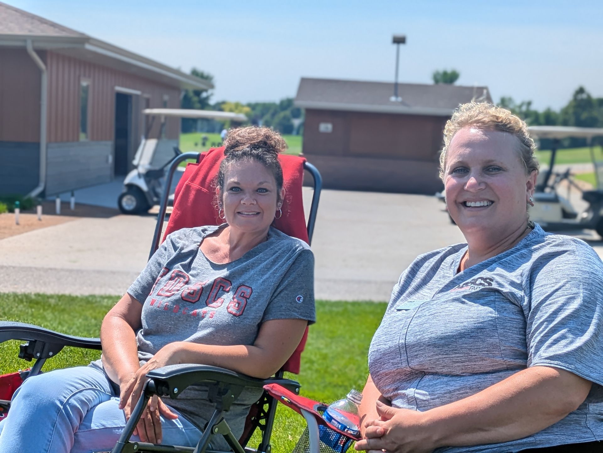Two women smiling in lawn chairs on a sunny day. Golf carts and a building are in the background.