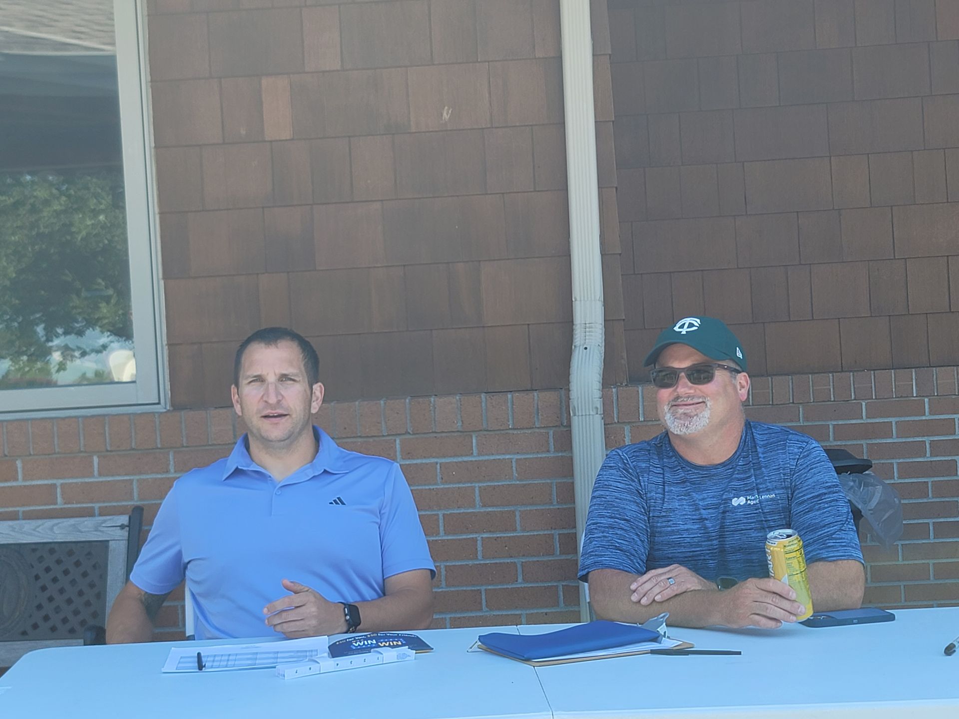 Two men at a table outside. One in a blue shirt, the other in a patterned shirt, holding a can. Both smiling.