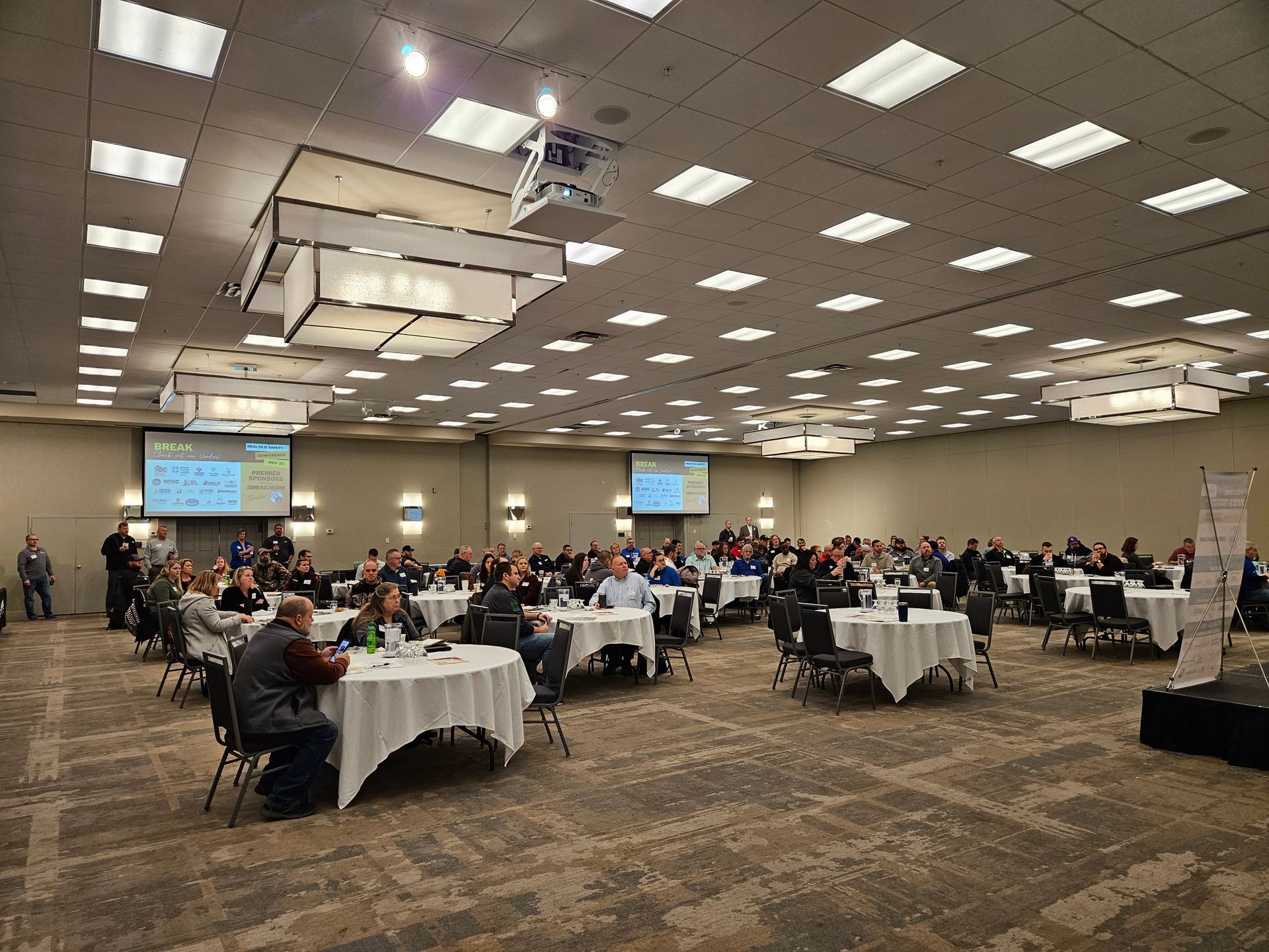Large event hall with round tables, attendees, and presentation screens.