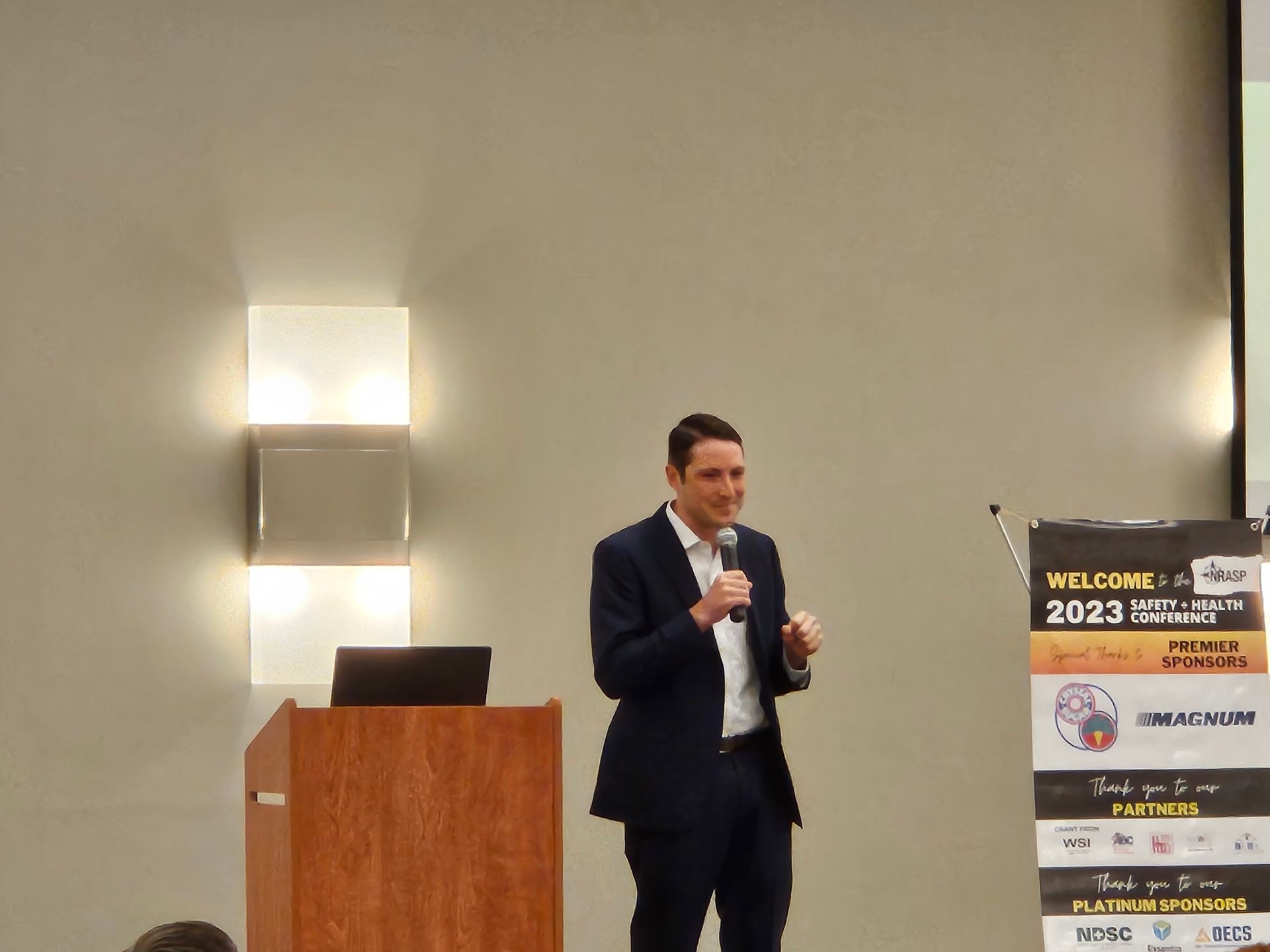 Man in suit speaking at a podium, gesturing. Conference banner and lighting in background.
