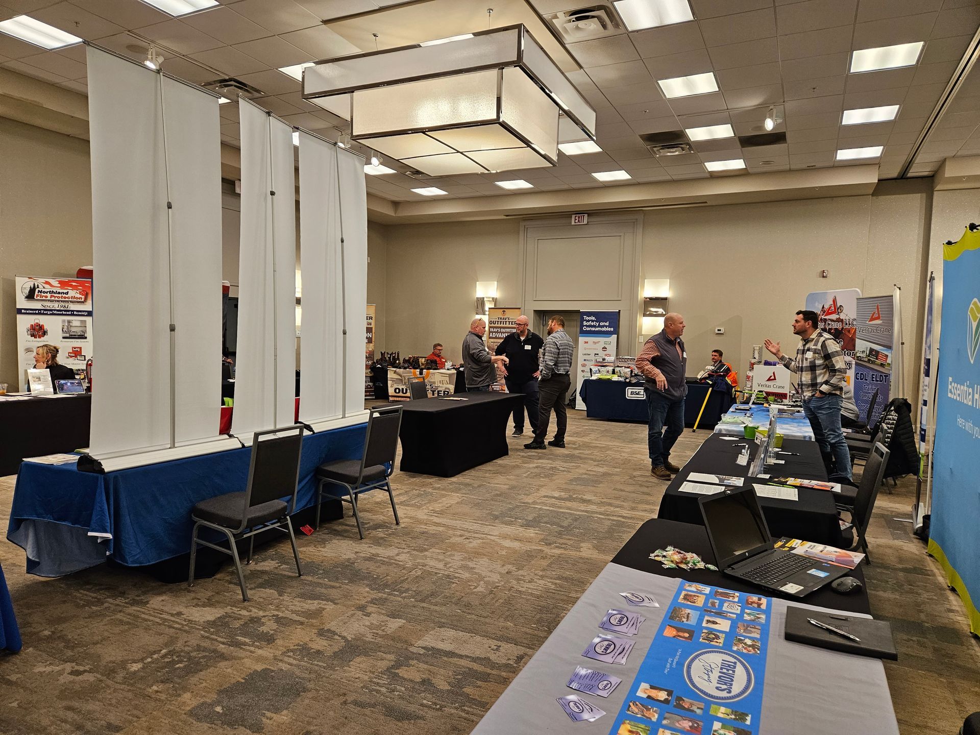 Trade show with booths, attendees conversing. Large room, various displays, tables, and people.