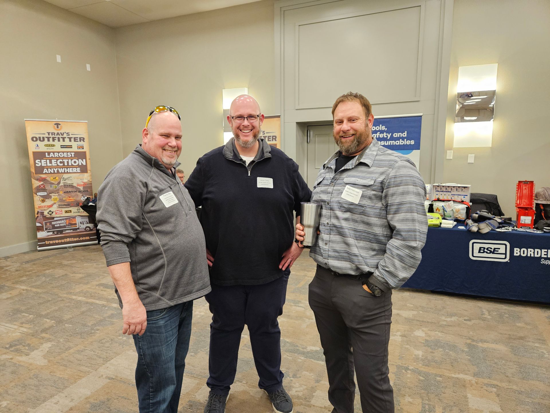 Three men stand together indoors at an event. They smile, wearing name tags and casual clothing.