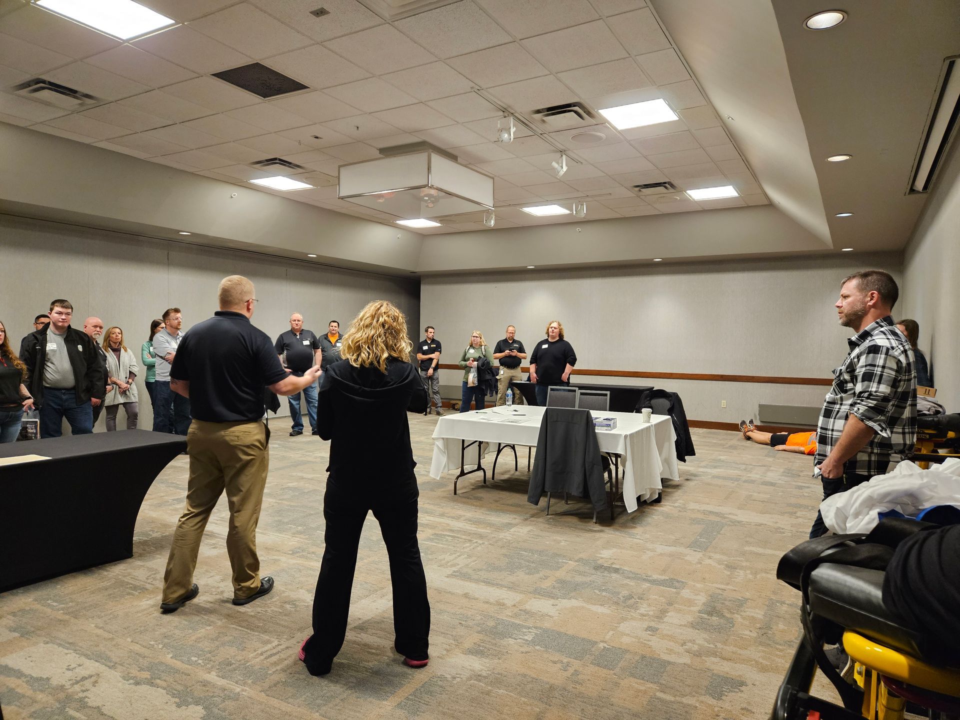 People in a training room, some standing, some at tables with equipment. Two people are in the foreground, facing the group.