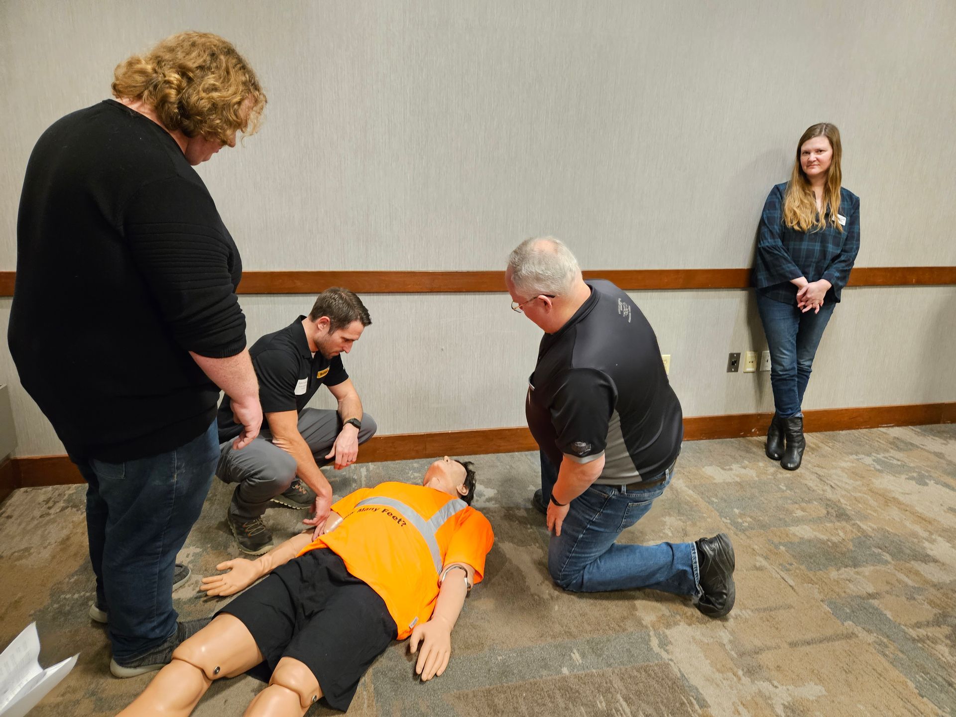 CPR training: Four people practice on a mannequin wearing an orange vest, one person watches.