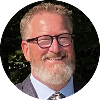 Man with glasses and beard smiles outdoors, wearing suit jacket and patterned tie.